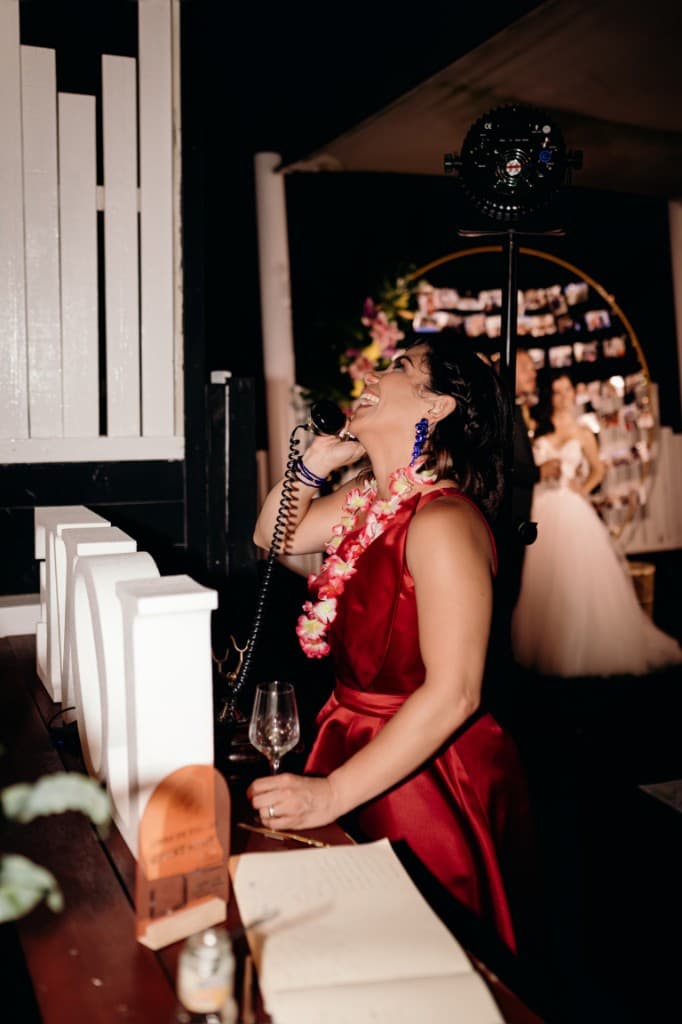 A guest in a red dress laughing while leaving a voice message on a vintage telephone at a wedding